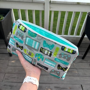 May include: A rectangular gray pouch with a teal interior and a zipper closure. The exterior features a pattern of video game controllers and consoles in teal, black, and lime green.