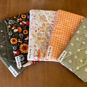 Puede incluir: Cinco artículos de tela acolchada con varios diseños, incluyendo setas, girasoles y margaritas. Las telas son en tonos verdes, naranjas y crema. Cada artículo tiene una etiqueta blanca que dice "Pixie & Thread".