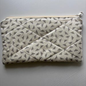 Op de afbeelding: Een crèmekleurige, gewatteerde etui met een herhaald patroon van kleine bijen. De etui heeft een beige rits en een diamantvormig stikselpatroon. De bijen zijn in een grijze kleur.