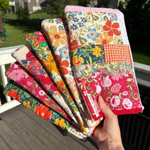May include: A collection of colorful, floral-patterned quilted fabric items, fanned out to display the variety of designs. The fabrics feature a mix of red, pink, orange, yellow, and blue flowers. The text "Pixie & Thread" is visible on a label.