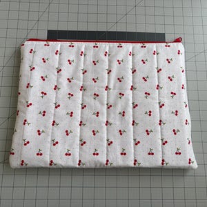 May include: White quilted fabric case with a red zipper and cherry pattern. The case is rectangular and has a red trim. The fabric has a repeating pattern of red cherries and small gray dots.