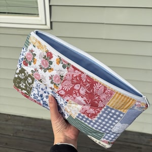 May include: A patchwork zippered pouch with a light blue interior. The exterior features a variety of floral and patterned fabrics in red, white, blue, and yellow. The zipper is white.