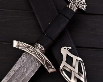 May include: A black leather-wrapped dagger with a silver blade and intricate metalwork on the handle. The blade has a damascus pattern and the handle has a bird design.