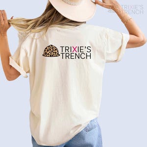 May include: A white t-shirt with a leopard print hard hat and the text "TRIXIE'S TRENCH" in black with a pink "X" in the word "TRIXIE'S".