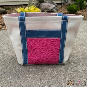 Puede incluir: Un bolso tote beige con un bolsillo con estampado de leopardo rosa y detalles en denim azul. El bolso tiene asas y ribetes de denim azul, con un forro interior rosa. El bolso está sobre una superficie de hormigón.