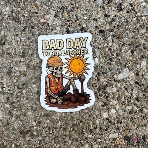 May include: A sticker with the text "BAD DAY TO BE A LABORER" above a skeleton wearing a hard hat and safety vest, holding a shovel and a sun. The sticker has a white border and is on a textured gray surface.