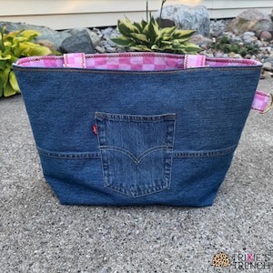 May include: A blue denim tote bag with a pink and white checkered interior lining. The bag features a back pocket and a red tag. The handles are pink and white striped. The bag is sitting on a gray surface.