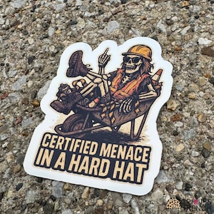 Puede incluir: Adhesivo con un esqueleto en una carretilla, con casco y gafas de sol, mostrando el dedo corazón. El texto dice "CERTIFIED MENACE IN A HARD HAT". Diseño de estilo vintage y desgastado.