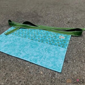 Puede incluir: Un delantal de tela turquesa y estampada con una correa verde. El delantal tiene una sección inferior azul claro y una sección superior estampada con pequeños diseños verdes. Las costuras blancas delinean las secciones y los bordes. El delantal está sobre una superficie gris.