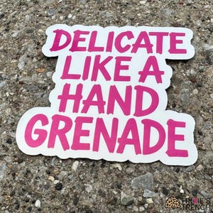 Puede incluir: Una pegatina blanca con borde rosa y el texto "DELICATE LIKE A HAND GRENADE" en rosa intenso. La pegatina está sobre una superficie gris texturizada.