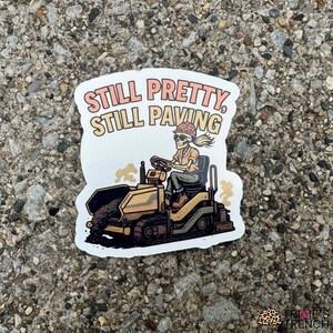 Puede incluir: Una pegatina que muestra un esqueleto operando una máquina pavimentadora. El esqueleto lleva un casco y ropa de trabajo. La pegatina tiene el texto "Still Pretty, Still Paving". La pegatina está sobre una superficie gris texturizada.