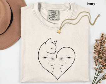 Può includere: T-shirt color avorio con un design a forma di gatto e cuore nero. Il gatto è racchiuso in un cuore pieno di stelle. Una collana con pendente a stella dorata è appoggiata sulla maglietta. La maglietta ha un'etichetta Comfort Colors ed è taglia XL-TG-EG.
