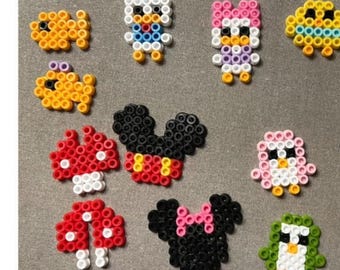 Può includere: Una collezione di 15 personaggi di pixel art colorati realizzati con perline Perler. I personaggi includono Topolino, un dinosauro rosa, un uccello rosa, un pulcino giallo, un fungo rosso, un pinguino blu e un pinguino verde.