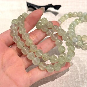 May include: A close-up of a hand holding two strands of light green prehnite gemstone bead bracelets. The beads are round and translucent, with subtle inclusions. A third strand of beads is visible in the background.