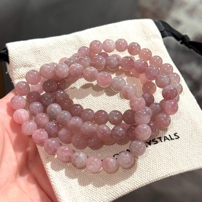 May include: Three strands of pink gemstone bead bracelets are displayed on a small, off-white canvas bag. The beads are a soft, translucent pink, and the bag has the word "CRYSTALS" printed on it in black. The bracelets are shown in a close-up shot.