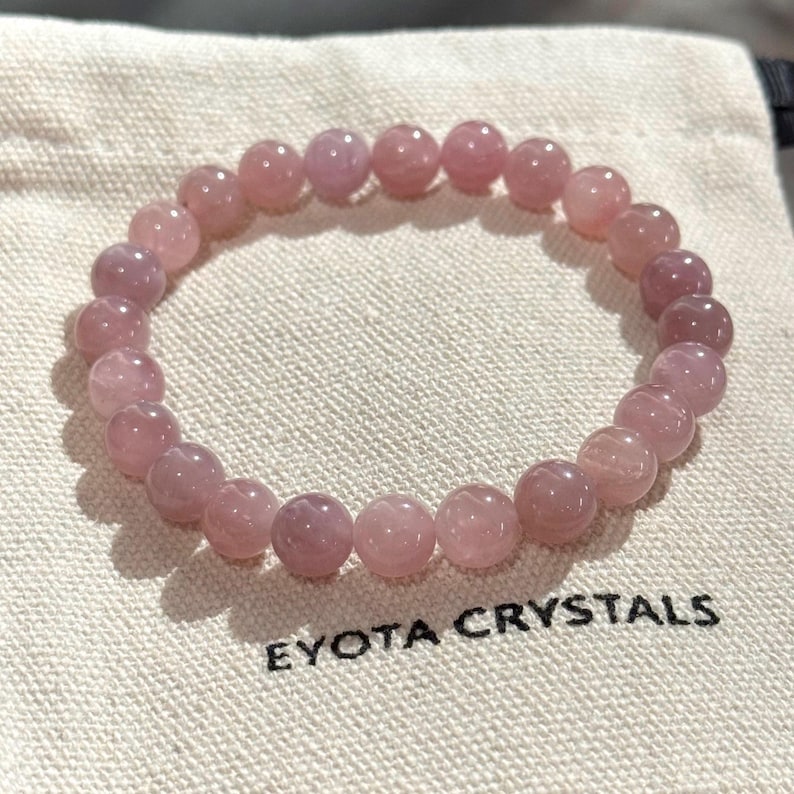 May include: A light pink gemstone bead bracelet. The bracelet is displayed on a beige cloth bag, with the text "EYOTA CRYSTALS" printed in black.