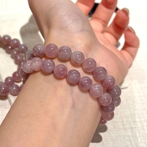 May include: Two bracelets of light purple, round gemstone beads are worn on a wrist. The beads have a translucent quality, and the bracelets are stacked. The skin tone is fair, and the background is neutral.