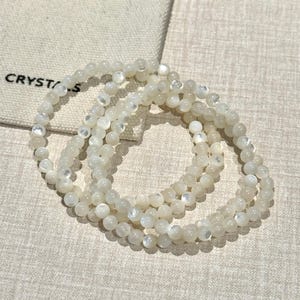 Puede incluir: Tres hilos de cuentas de piedras preciosas blancas e iridiscentes están dispuestas sobre una superficie de tono neutro. Las cuentas son pequeñas y redondas, con un brillo sutil. Una pequeña bolsa blanquecina con la palabra "CRYSTALS" es visible en el fondo.