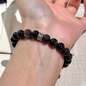 Könnte beinhalten: Ein Armband aus schwarzem Obsidian mit einer silbernen Zwischenperle. Das Armband besteht aus runden, polierten schwarzen Perlen. Die silberne Perle hat ein dekoratives Design. Das Armband wird am Handgelenk getragen.