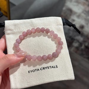 May include: A light pink beaded bracelet sits on a small, off-white pouch. The bracelet is made of round, polished beads. The pouch has the words "EYOTA CRYSTALS" printed in black. The bracelet is held by a person.