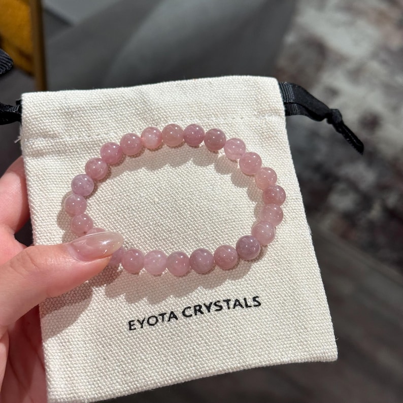 May include: A light pink beaded bracelet sits on a small, off-white pouch. The bracelet is made of round, polished beads. The pouch has the words "EYOTA CRYSTALS" printed in black. The bracelet is held by a person.