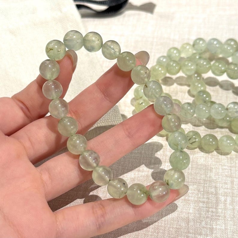 May include: A light green beaded bracelet with round beads. The bracelet is made of translucent, pale green stones. The beads are strung together to form a circular shape. Another bracelet is visible in the background.