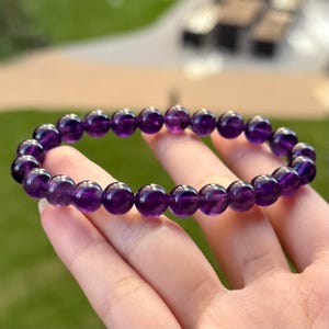 Op de afbeelding: Een armband met gepolijste, ronde, dieppaarse amethiststenen. De kralen zijn aan elkaar geregen, waarbij de natuurlijke kleur en textuur van de edelstenen worden getoond. De armband is een sieraad.