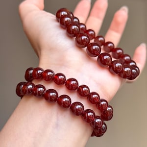 Puede incluir: Una pulsera elástica compuesta por cuentas redondas y lisas de granate rojo intenso. Las cuentas presentan una superficie pulida y reflectante que capta la luz, creando un color carmesí oscuro y uniforme en todo el hilo de cuentas de piedra.