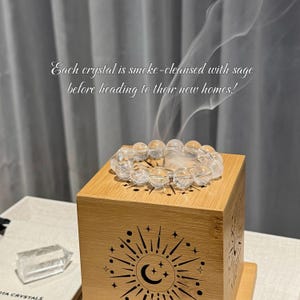 May include: A clear quartz crystal bracelet sits on a wooden incense holder, with smoke rising. The holder features a sun and moon design. Text reads: "Each crystal is smoke-cleansed with sage before heading to their new homes!"