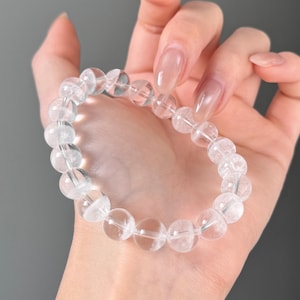 Könnte beinhalten: Ein Armband aus klaren Quarz-Kristallperlen. Das Armband besteht aus runden, durchscheinenden Perlen, die aneinandergereiht sind. Die Perlen haben ein leicht trübes Aussehen und werden von einem transparenten Gummiband zusammengehalten. Das Armband wird auf einer Hand gezeigt.