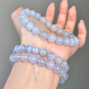 May include: Two light blue beaded bracelets. Each bracelet is made of round, polished beads. The bracelets are held in a hand, with the beads appearing to be approximately 1 cm in diameter. The bracelets have a soft, translucent appearance.