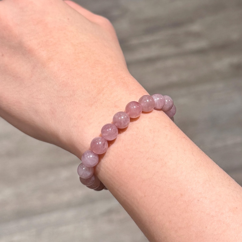 May include: A light purple beaded bracelet. The bracelet features round, polished beads in shades of lavender. The beads are strung together to form a bracelet. The bracelet is worn on a wrist.