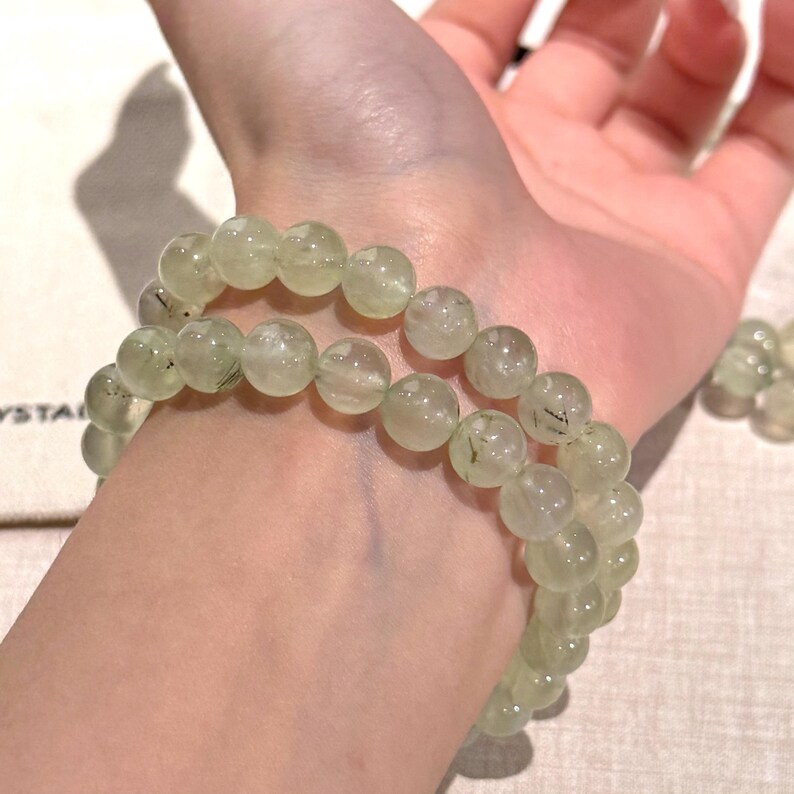 May include: Two light green beaded bracelets with translucent beads are worn on a wrist. The beads have a speckled appearance. The bracelets are made of round beads strung together. The background is a neutral color.