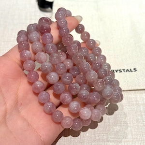 May include: Several bracelets composed of round, pale purple beads. The beads appear slightly translucent. The bracelets are held in a hand, with the word "RYSTALS" visible on a white bag in the background.