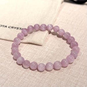 May include: A light purple beaded bracelet. The bracelet is made of round, polished beads. The beads are strung together on an elastic band. The bracelet is resting on a beige surface next to a small bag with the text "OTA CRYST" printed on it.