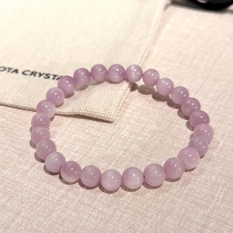 May include: A light purple beaded bracelet. The bracelet is made of round, polished beads. The beads are strung together on an elastic band. The bracelet is resting on a beige surface next to a small bag with the text "OTA CRYST" printed on it.