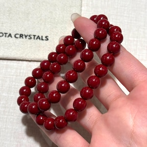 Op de afbeelding: Twee strengen dieprode kralenarmbanden. De ronde kralen hebben een glanzende afwerking. De armbanden worden in een hand gehouden, met een witte stoffen tas op de achtergrond met de tekst "OTA CRYSTALS".