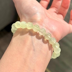 May include: A light green beaded bracelet made of round, translucent stones. The bracelet is worn on a wrist, showcasing the natural color and texture of the gemstones. The beads are approximately 0.5 inches in diameter.