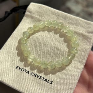 May include: A light green beaded bracelet rests on a beige pouch. The bracelet is made of round, translucent beads. The pouch has the text "EYOTA CRYSTALS" printed on it in black. The bracelet is a piece of jewelry.