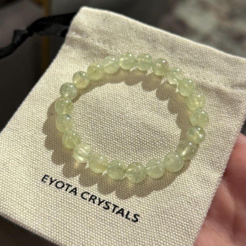 May include: A light green beaded bracelet rests on a beige pouch. The bracelet is made of round, translucent beads. The pouch has the text "EYOTA CRYSTALS" printed on it in black. The bracelet is a piece of jewelry.