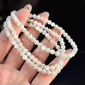 May include: A multi-strand bracelet composed of small, round, iridescent white beads with a smooth, polished finish. The beads exhibit a subtle pearlescent lustre, reflecting light across their spherical surfaces, arranged in a continuous circular loop.