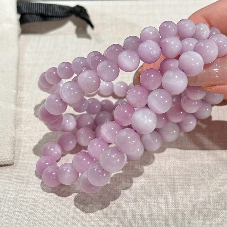 May include: Three light purple beaded bracelets. Each bracelet is made of round, polished beads. The bracelets are held together, showing the elastic band. The beads are a soft, pastel purple color. The bracelets are displayed on a light-colored surface.