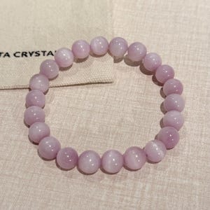 May include: A light purple beaded bracelet. The bracelet is made of round, polished beads. The beads are strung together on an elastic band. The bracelet is displayed on a neutral-colored surface.