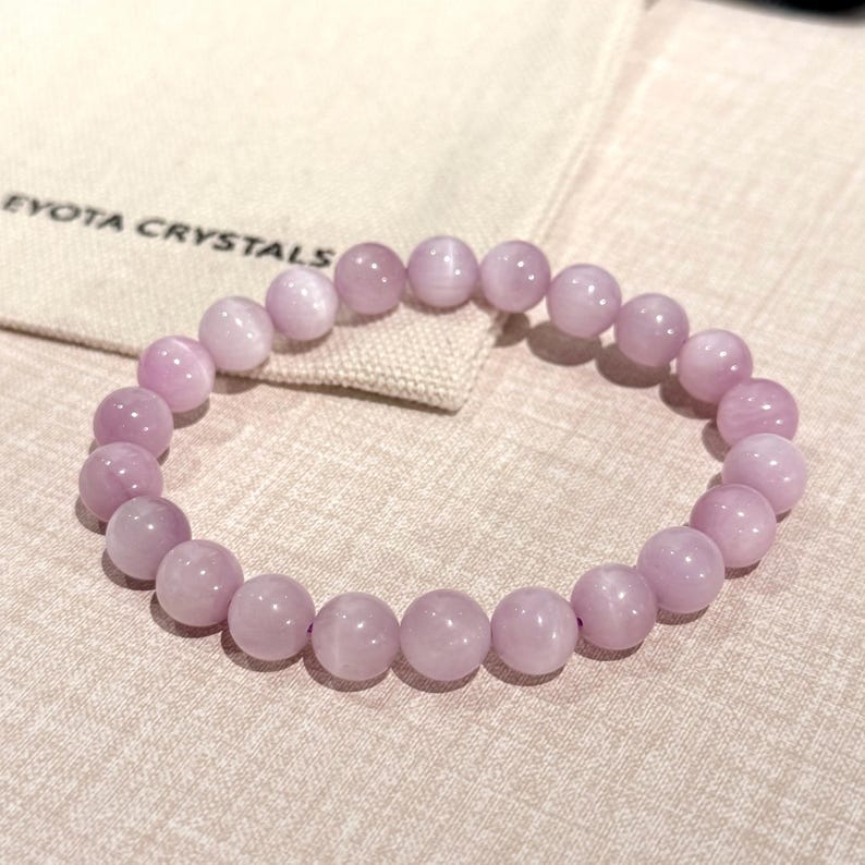 May include: A light purple beaded bracelet with round beads. The bracelet is resting on a beige cloth with the text "EYOTA CRYSTALS" printed on it. The beads are approximately 0.8 cm in diameter.