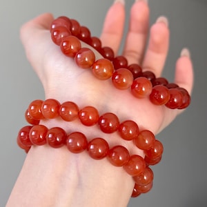 May include: Two strands of red-orange gemstone bead bracelets. The round, polished beads have a glossy finish. The bracelets are shown against a neutral background, emphasising the vibrant colour and texture. These bracelets are a type of jewellery.