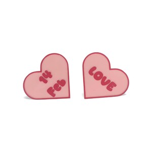 Pode incluir: Duas decorações em forma de coração rosa. Um coração tem o texto "14 Feb" em rosa mais escuro, e o outro diz "LOVE". Os itens são provavelmente para o Dia dos Namorados.