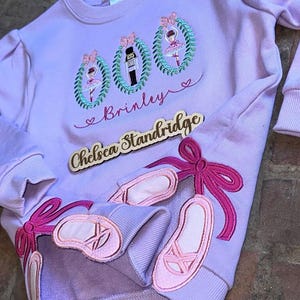May include: A lilac-colored sweatshirt featuring ballet-themed embroidery and appliqués. The design includes three wreath-framed ballerinas and a nutcracker, with the name "Brinley" and ballet slippers. A wooden name tag says "Chelsea Standridge".