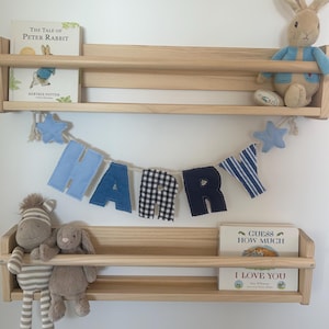 May include: Two light-coloured wooden wall shelves holding a book titled The Tale of Peter Rabbit, a plush rabbit, a zebra plush, a rabbit plush, and a book titled Guess How Much I Love You. A fabric banner spells the name HARRY in blue and black.