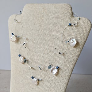 May include: A delicate necklace made with silver wire, featuring a series of white baroque pearls and dark blue beads. The necklace is displayed on a beige fabric mannequin.