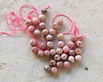May include: A collection of pink and white patterned beads strung on pink thread. The beads are round and have a natural, polished look.